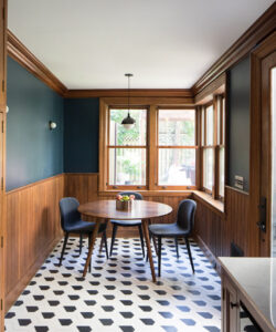 Modern dining nook in Brooklyn brownstone with deep blue walls, warm wooden trim, geometric tile flooring, and round wooden table accompanied by sleek chairs.