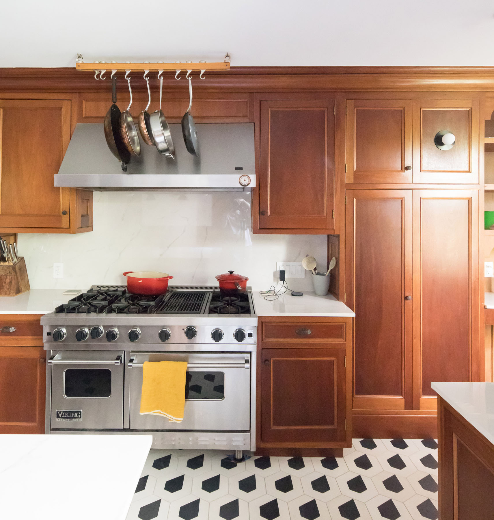 Elegant modern kitchen renovation showcasing rich wooden cabinetry, a professional Viking stove, hanging stainless steel cookware, marble backsplash, and stylish geometric floor tiles.