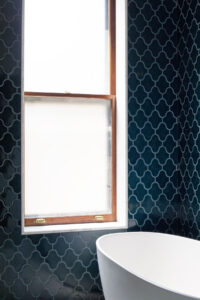 Stylish bathroom corner highlighting deep teal tiles with a distinctive wave pattern, paired with natural wooden-framed frosted windows, and complemented by a sleek white freestanding bathtub that exudes modern elegance.