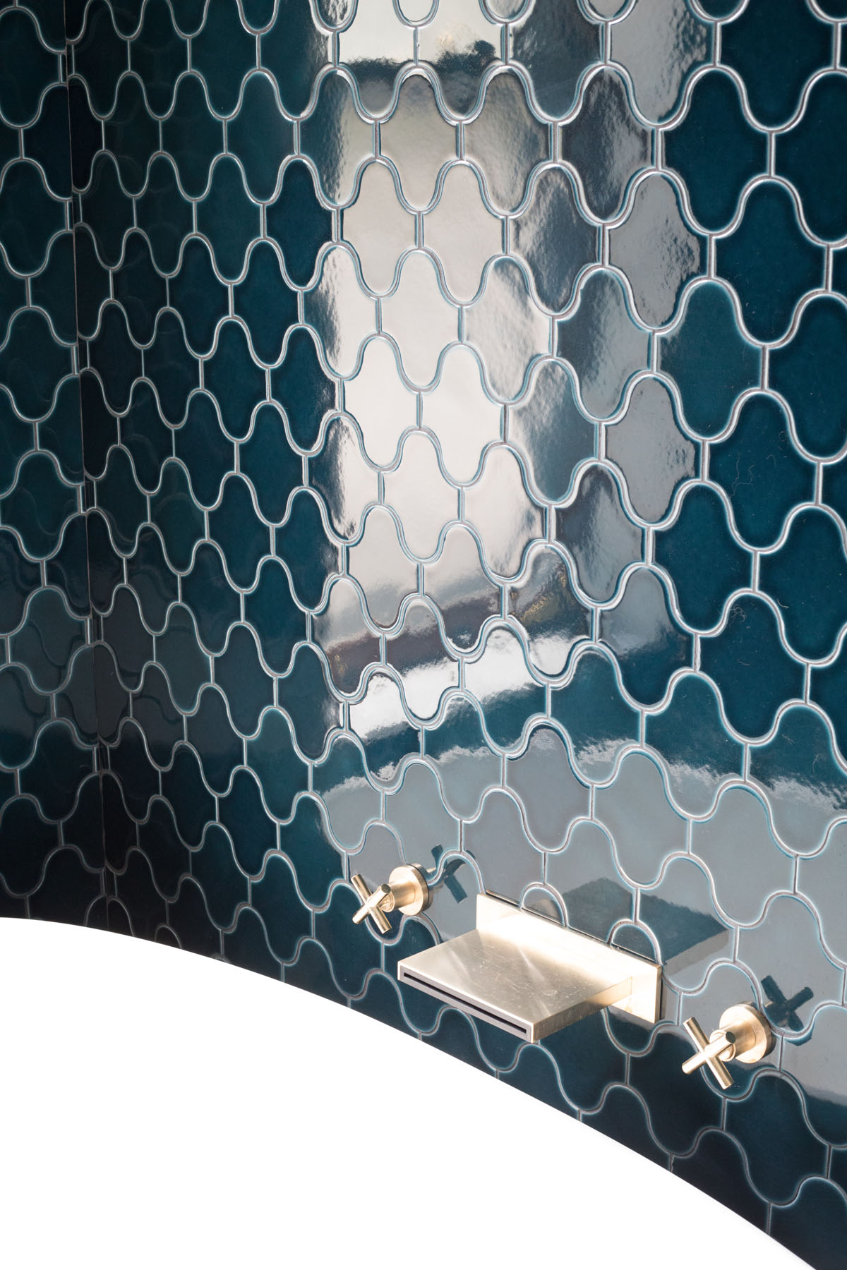 Detail of a contemporary bathroom featuring glossy deep blue clover-shaped tiles with light reflections, complemented by a sleek gold-colored faucet and fixture against a white backdrop.