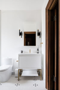 Elegant minimalist bathroom featuring a pristine white vanity with golden accents, modern white toilet, chic dual black and gold wall sconces, a large square mirror with a wooden frame, and a detailed wooden door with vintage latch.