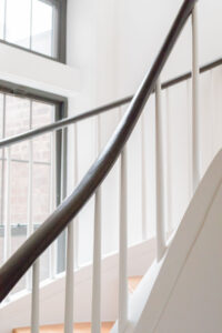 Close-up of a dark-stained handrail with a smooth, oval profile, ascending alongside white balusters and a white stairway against a backdrop of bright windows in a New York City townhouse renovation by Rauch Architecture.