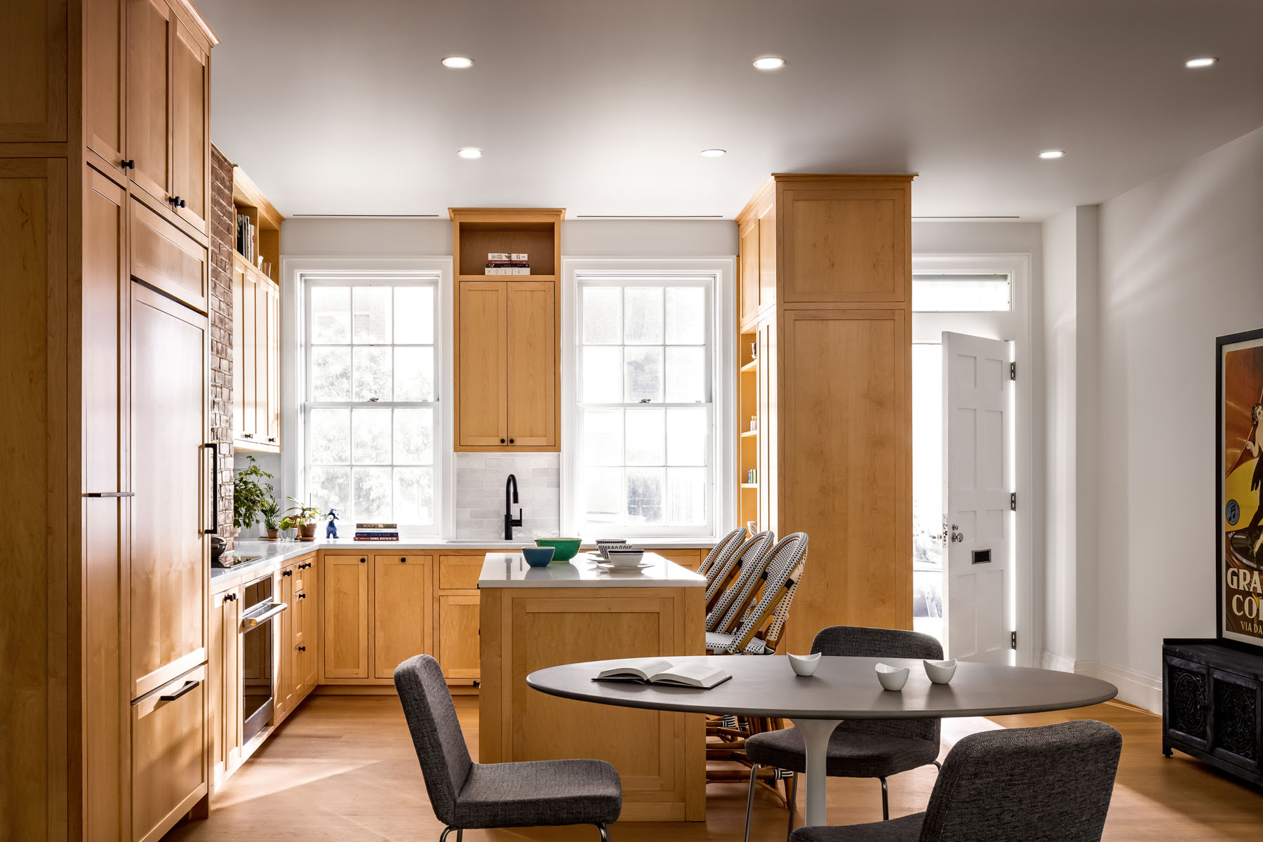 Spacious kitchen interior featuring natural wood cabinetry, a brick backsplash, large windows letting in natural light, and a cozy dining area with a round table, gray chairs, and a contemporary art piece on the wall.