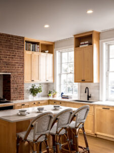 Rauch Architecture remodels a cozy kitchen in Tribeca that blends modern and rustic styles with tall maple cabinets, alcove transoms displaying cookbooks, ceiling lights emitting a soft glow, and a standout exposed brick wall.
