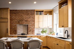 Modern kitchen interior with exposed brick wall, natural wooden cabinetry, white subway tile backsplash, and an intimate dining area with woven chairs.