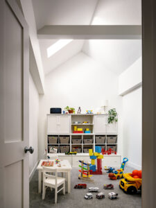 Bright and spacious children's playroom in historic Tribeca townhouse on NYC's Harrison St. with architectural skylight detailing, custom white storage cabinetry, and an array of colorful toys and games on a soft gray carpet.