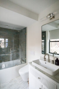Modernized Upper East Side bathroom featuring glass-enclosed shower with gray subway tiles, white freestanding sink on a white vanity with marble countertop, and herringbone-patterned tile floor