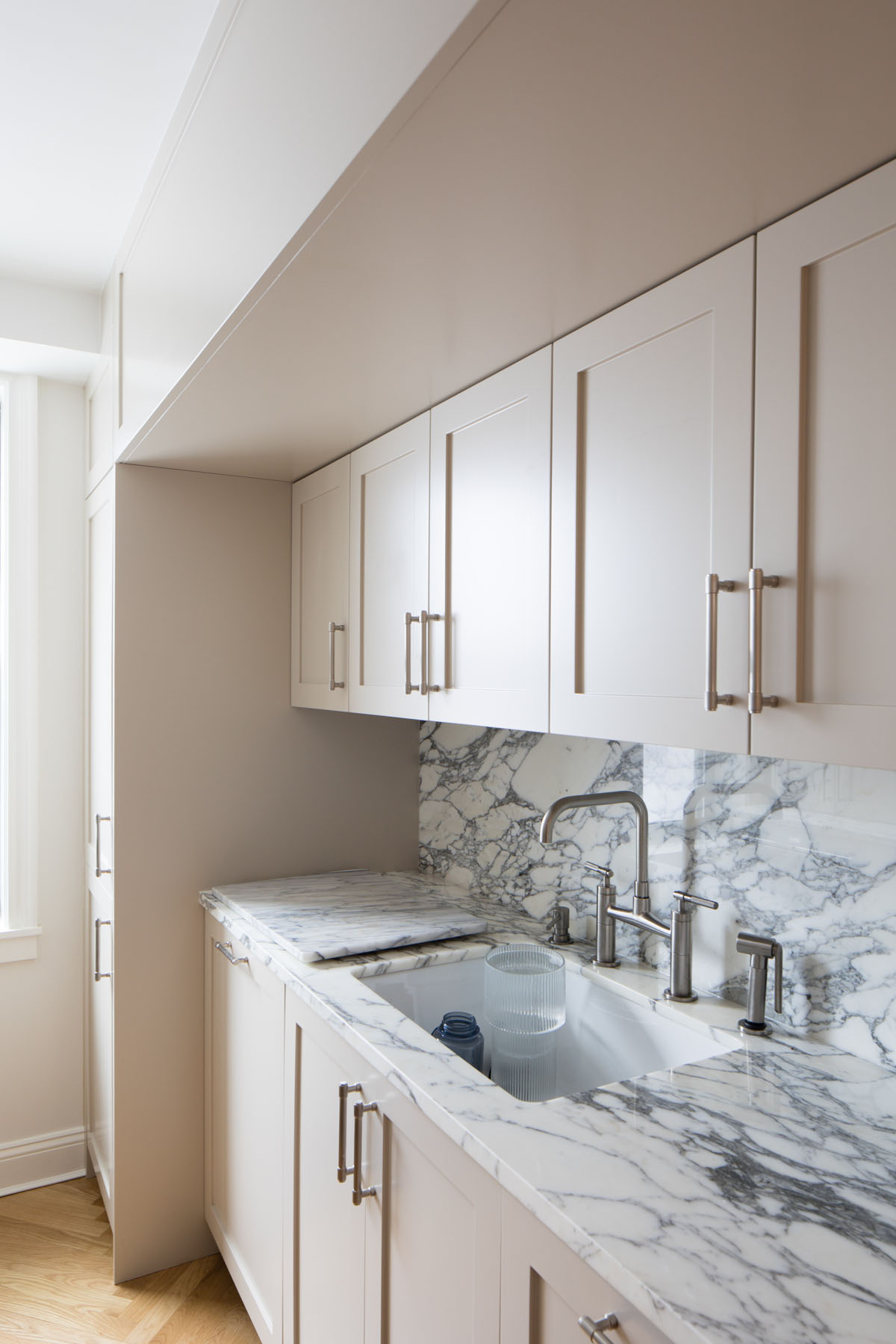 Upper East Side kitchen corner with marble splashback, white sink, beige cabinetry, and herringbone wood floor designed by Rauch Architecture.