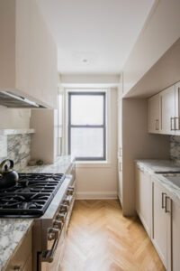 Newly renovated kitchen in Upper East Side co-op featuring a stainless steel gas stove, marble countertops, white cabinetry, and herringbone wood floors by Rauch Architecture