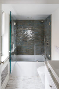 Upper East Side bathroom with glass-enclosed shower, subway tile walls in shades of gray, white porcelain toilet, and a herringbone pattern floor.