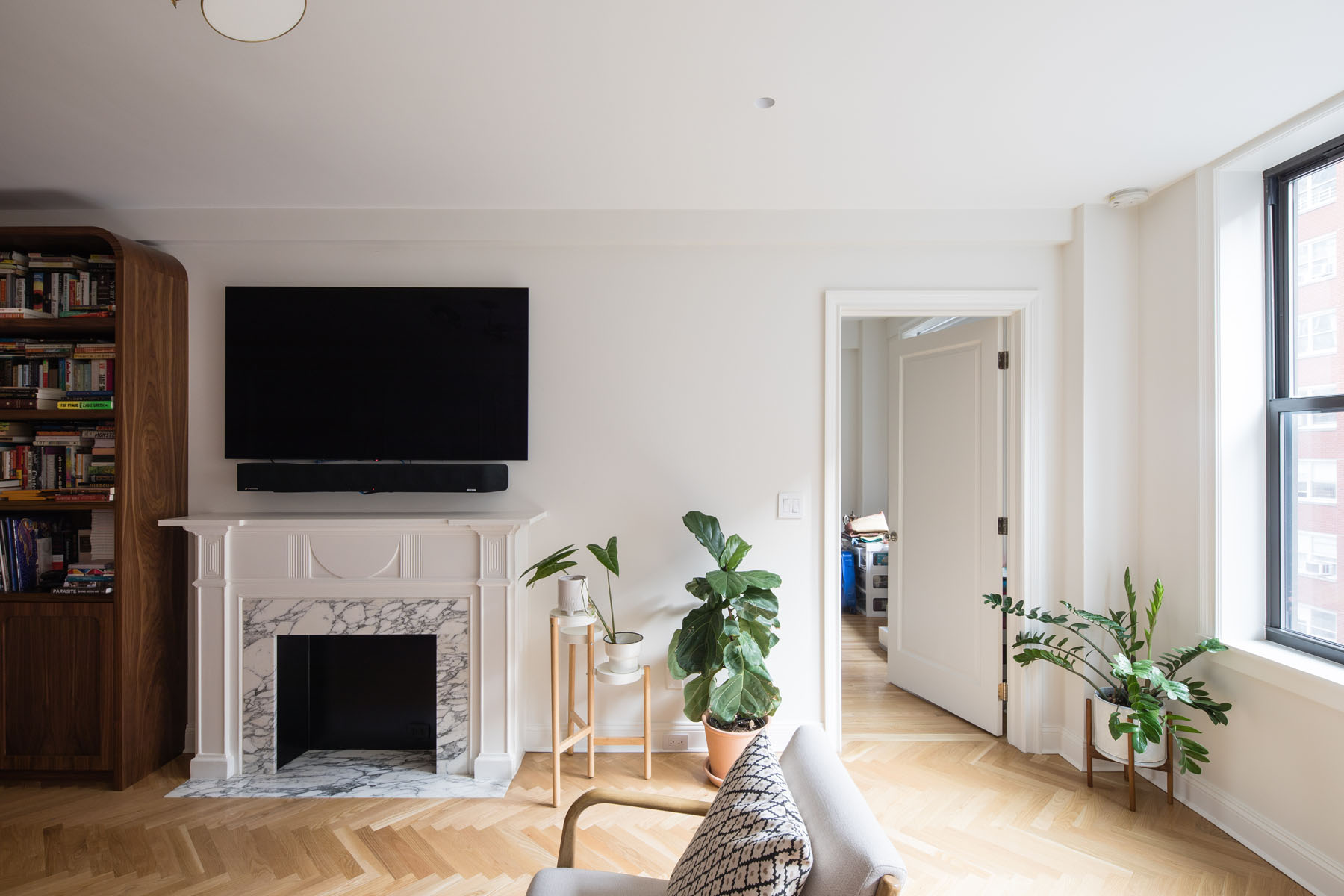Upper East Side living room with traditional fireplace, mounted flat-screen TV, herringbone wood flooring, and potted green plants, designed by Rauch Architecture.