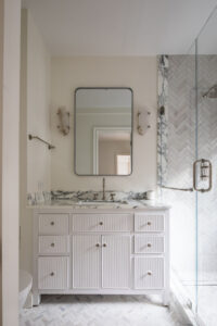 New York City co-op bathroom with herringbone marble tile, glass shower, white beadboard vanity with marble top, and rectangular mirror with wall-mounted lights.