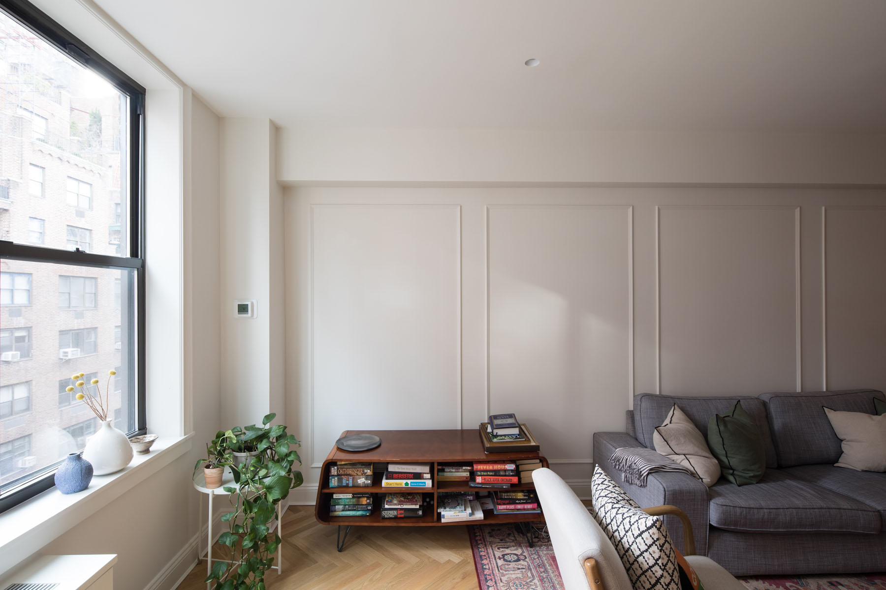 Bright Upper East Side living room with large window, gray sofa, wooden coffee table with books, and potted plants against a backdrop of paneled walls by Rauch Architecture.