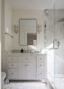 New York City co-op bathroom with herringbone marble tile, glass shower, white beadboard vanity with marble top, and rectangular mirror with wall-mounted lights.