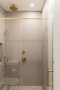 Modern bathroom with gray tiles, gold fixtures, and a glass shower enclosure in a Chelsea, NYC duplex by Rauch Architecture.